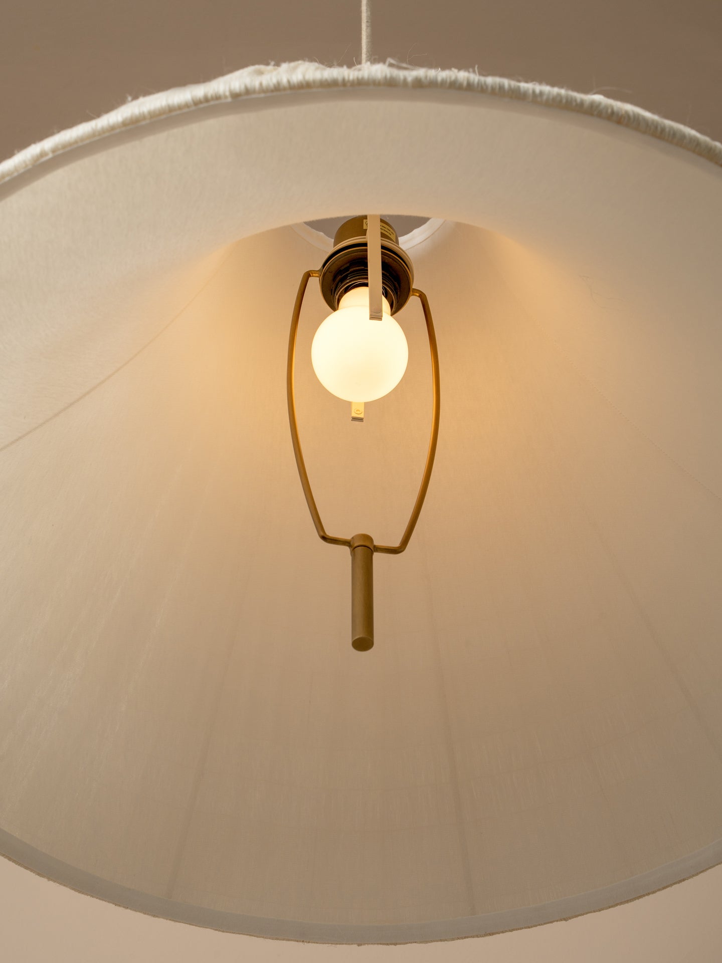 A close-up of a hanging pendant light with a large cream fabric drum shade, a warm white globe bulb, and a brass-toned metal frame against a neutral beige background.