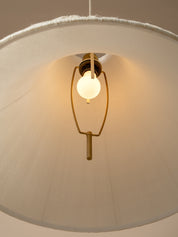 A close-up of a hanging pendant light with a large cream fabric drum shade, a warm white globe bulb, and a brass-toned metal frame against a neutral beige background.