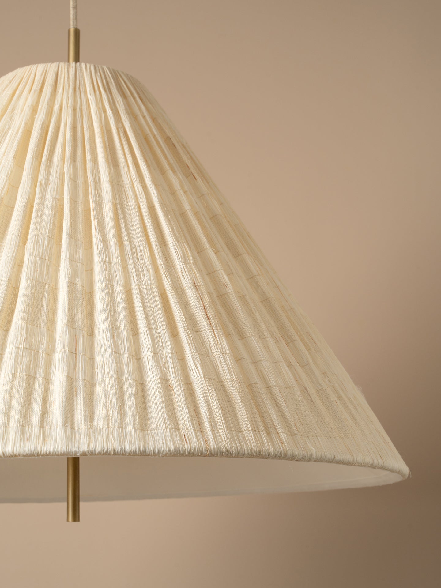 A close-up of a pleated conical pendant lampshade in a warm cream or off-white tone, suspended by slim brass-toned metal rods against a solid beige background.