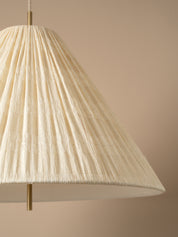 A close-up of a pleated conical pendant lampshade in a warm cream or off-white tone, suspended by slim brass-toned metal rods against a solid beige background.