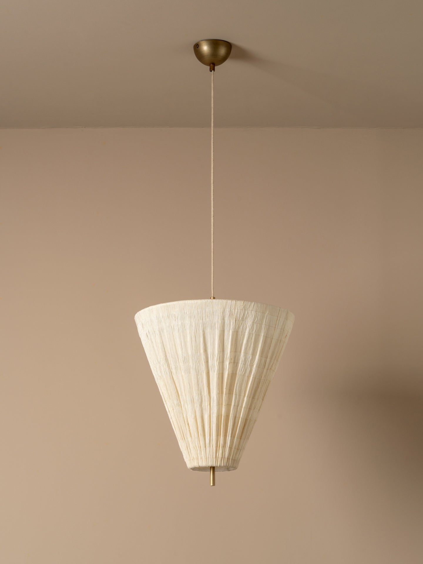 A modern pendant light with a pleated off-white paper cone shade and a brass ceiling canopy, shown hanging against a warm beige wall.