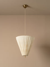 A modern pendant light with a pleated off-white paper cone shade and a brass ceiling canopy, shown hanging against a warm beige wall.