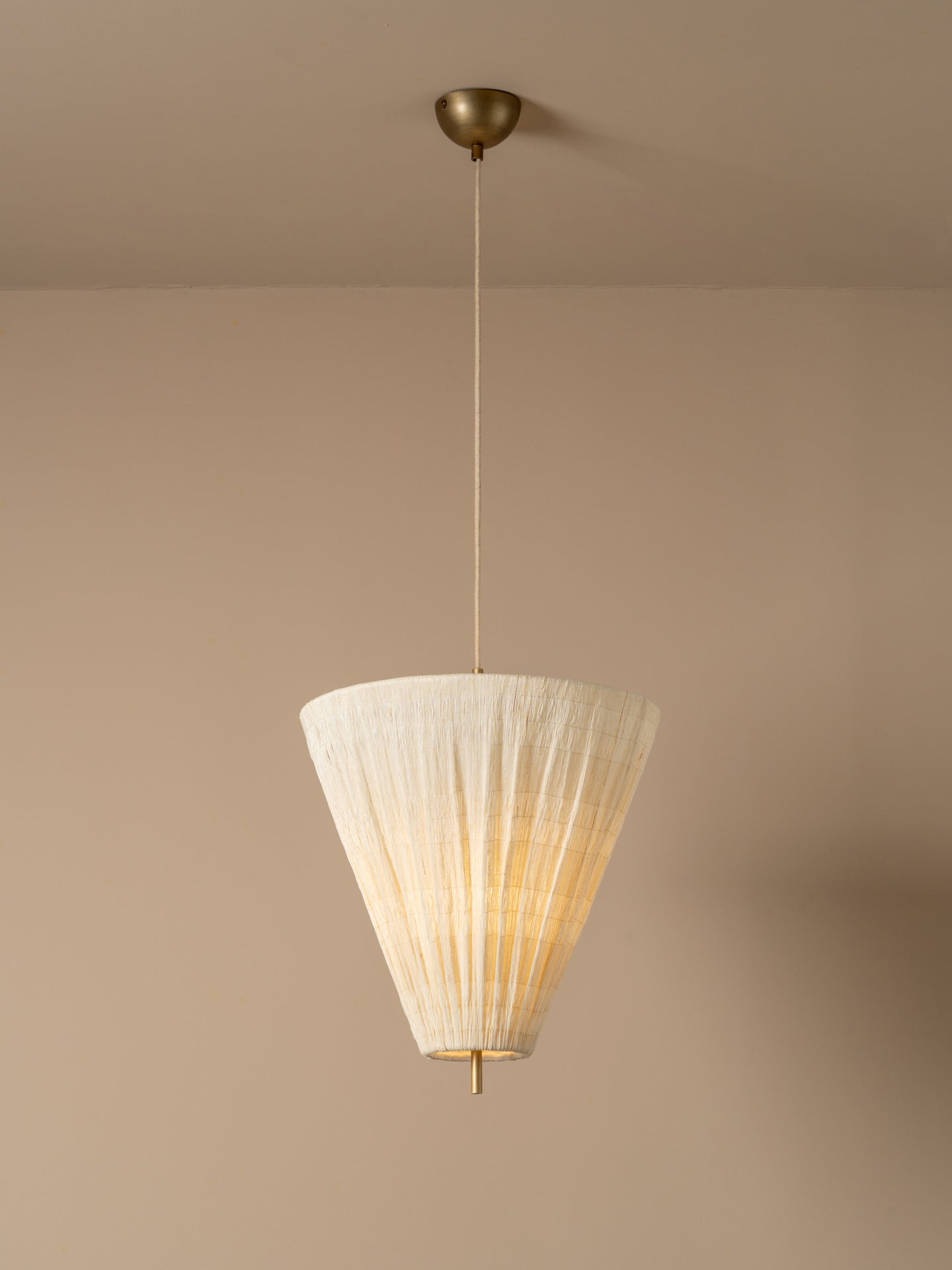 A modern pendant light illuminated with a pleated off-white paper cone shade and a brass ceiling canopy, shown hanging against a warm beige wall.