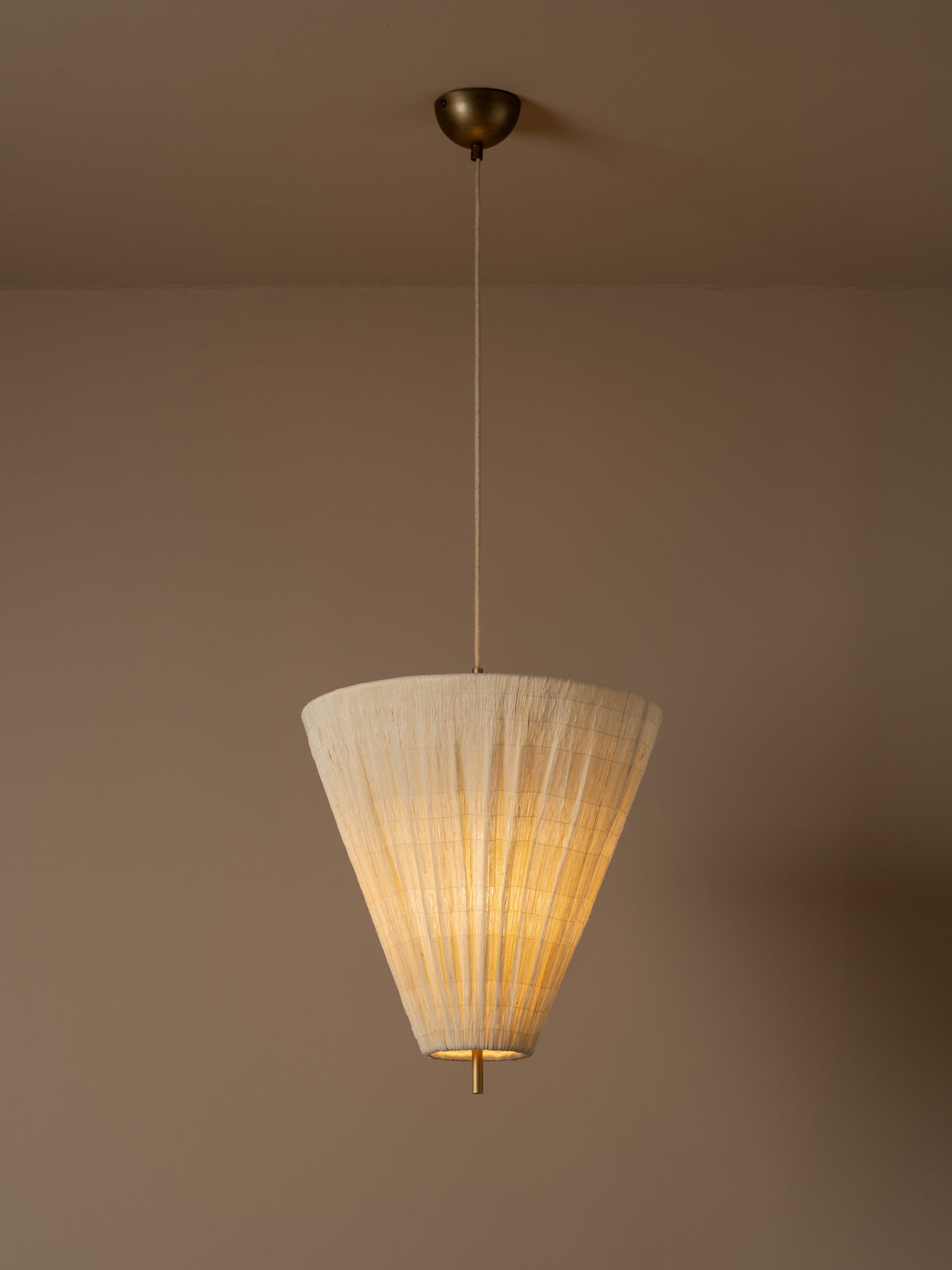 A modern pendant light illuminated with a pleated off-white paper cone shade and a brass ceiling canopy, shown hanging against a warm beige wall.