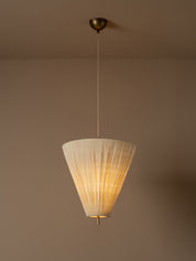 A modern pendant light illuminated with a pleated off-white paper cone shade and a brass ceiling canopy, shown hanging against a warm beige wall.