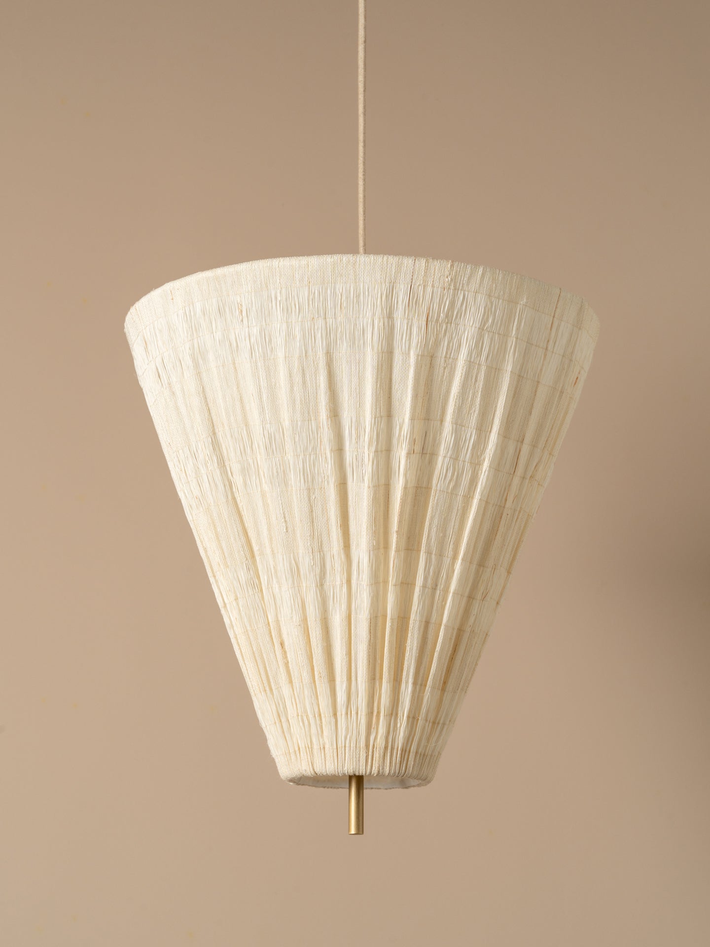 A modern pendant light  with a pleated off-white paper cone shade and a brass ceiling canopy, shown hanging against a warm beige wall.