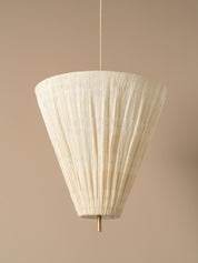 A modern pendant light  with a pleated off-white paper cone shade and a brass ceiling canopy, shown hanging against a warm beige wall.