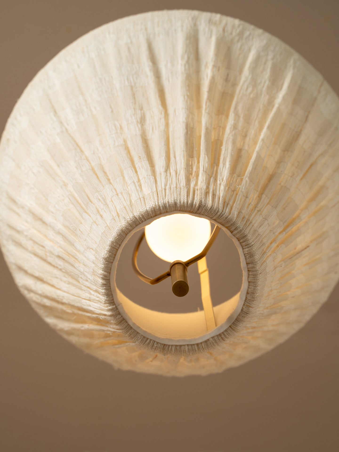 A close-up of a warm white textured fabric drum lampshade with a visible brass socket and glowing bulb inside, set against a solid warm beige background.