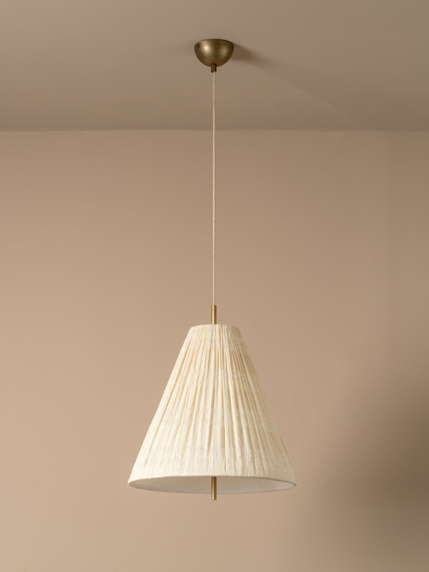 A modern pendant light with a pleated off-white paper cone shade and a brass ceiling canopy, shown hanging against a warm beige wall.