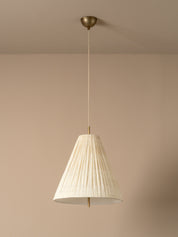 A modern pendant light with a pleated off-white paper cone shade and a brass ceiling canopy, shown hanging against a warm beige wall.