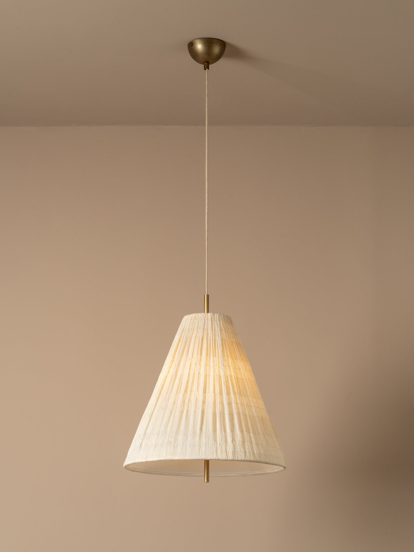 A close-up of a warm white textured fabric drum lampshade with a visible brass socket and glowing bulb inside, set against a solid warm beige background.