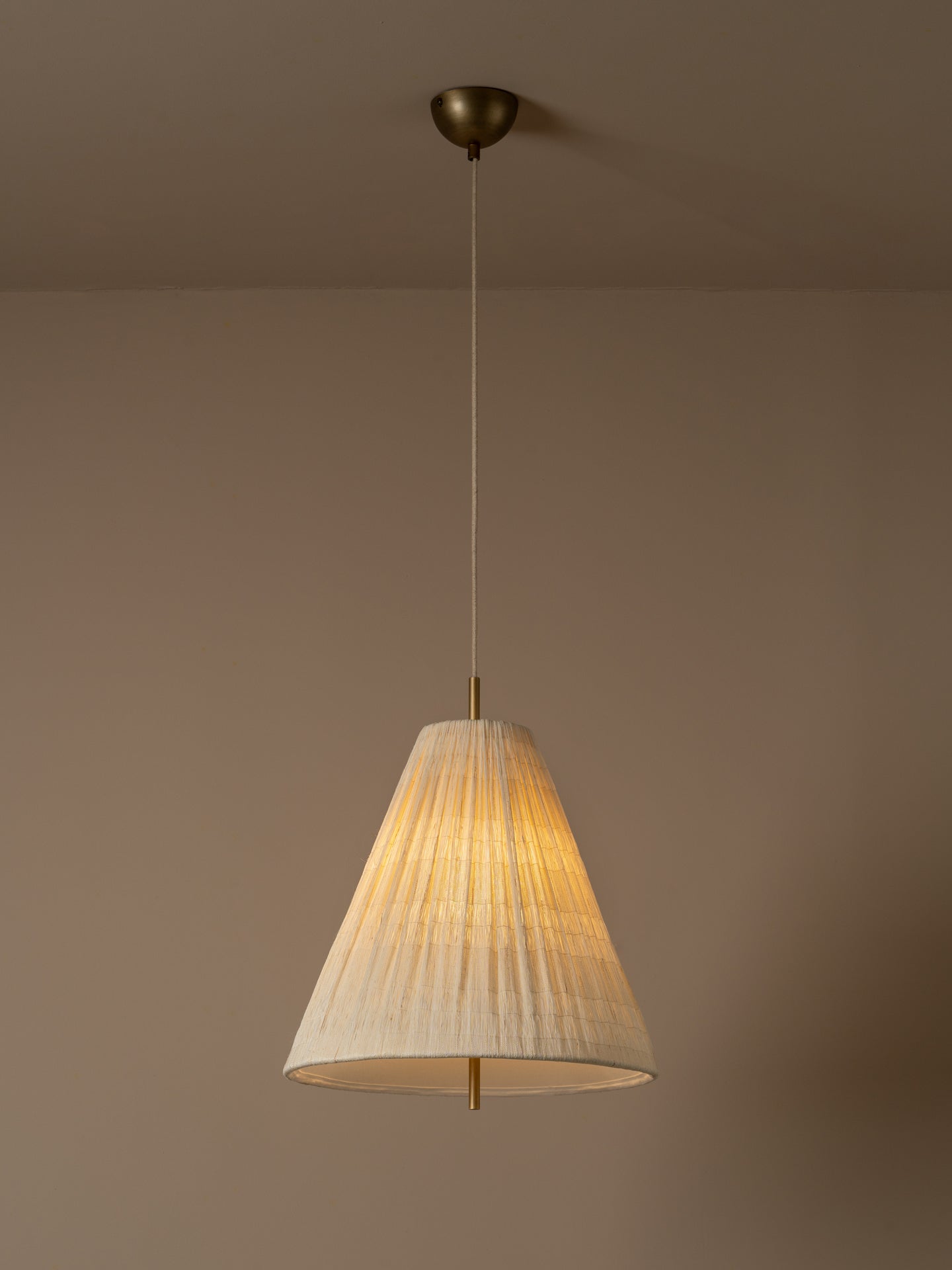 A conical pendant light shade made of textured, pleated cream-colored paper or fabric, suspended by a thin cord against a solid warm beige background. light is on.