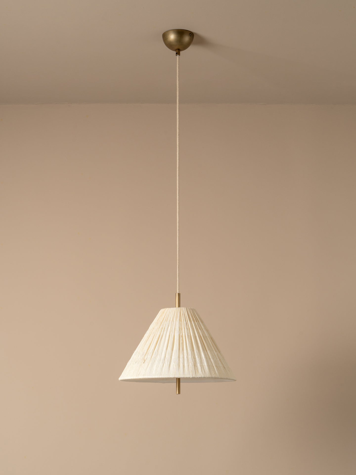 A modern pendant light with a pleated white conical shade and a brass ceiling mount, suspended against a minimalist beige wall and ceiling.