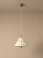 A modern pendant light with a pleated white conical shade and a brass ceiling mount, suspended against a minimalist beige wall and ceiling.