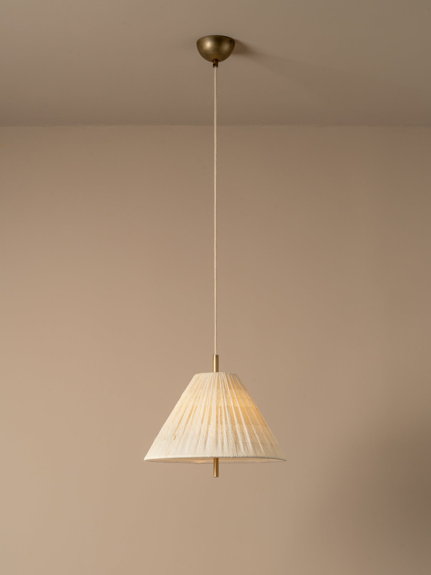A modern pendant light illuminated with a pleated white conical shade and a brass ceiling mount, suspended against a minimalist beige wall and ceiling.