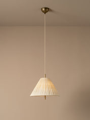 A modern pendant light illuminated with a pleated white conical shade and a brass ceiling mount, suspended against a minimalist beige wall and ceiling.