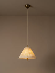 A modern pendant light illuminated with a pleated white conical shade and a brass ceiling mount, suspended against a minimalist beige wall and ceiling.