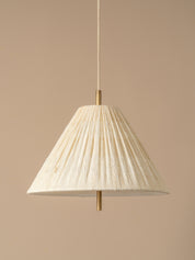 A modern pendant light with a pleated white conical shade and a brass ceiling mount, suspended against a minimalist beige wall and ceiling.