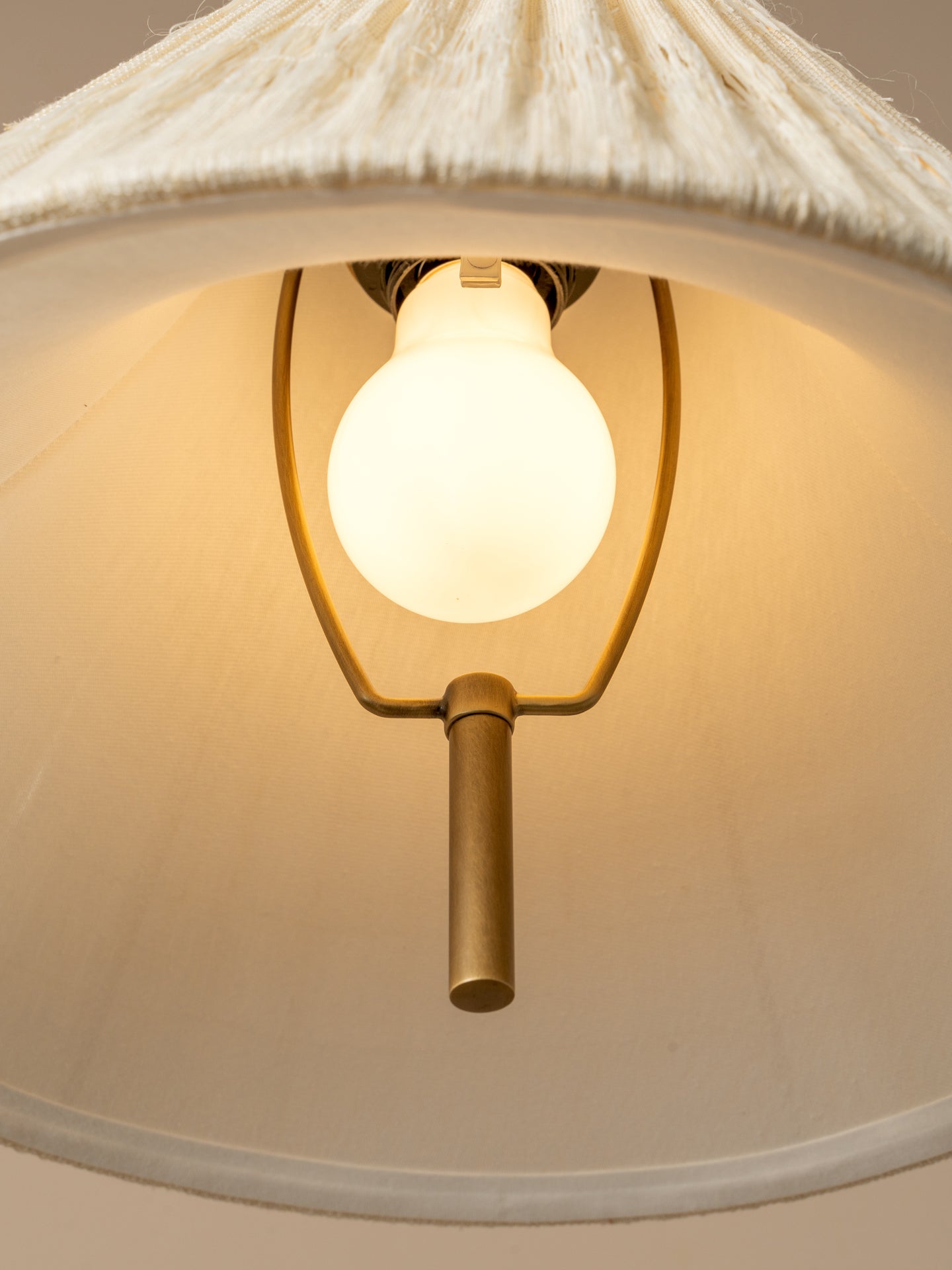 A close-up of a hanging pendant light with a large white fabric drum shade, a glowing warm white globe bulb, and a brass-toned metal frame against a neutral beige background.