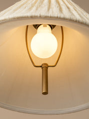 A close-up of a hanging pendant light with a large white fabric drum shade, a glowing warm white globe bulb, and a brass-toned metal frame against a neutral beige background.