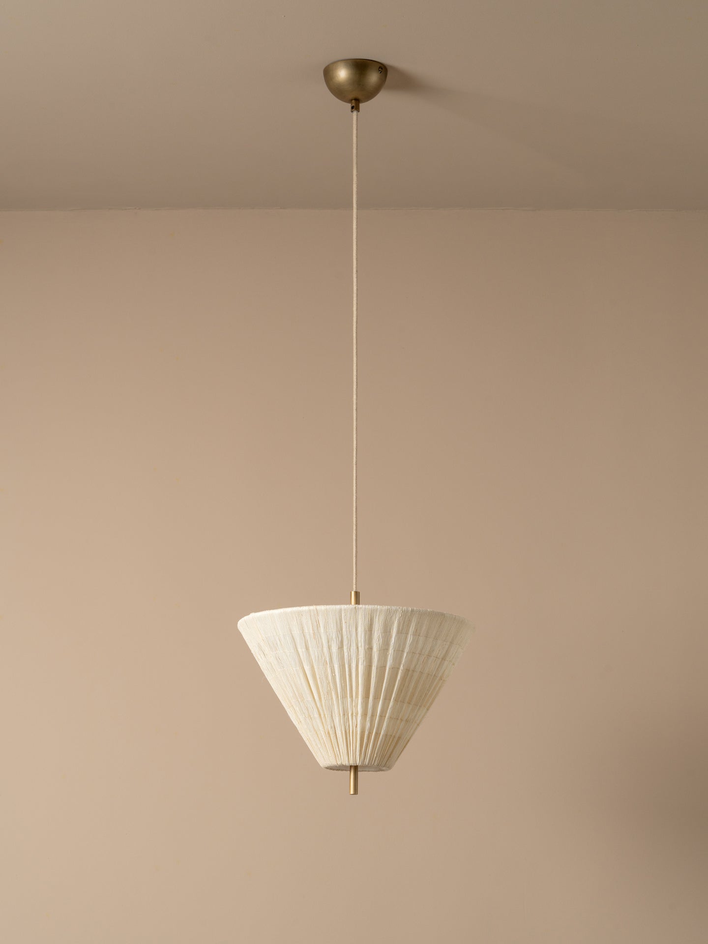 A modern pendant light with a pleated white conical shade and a brass ceiling mount, suspended against a minimalist beige wall and ceiling.