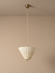 A modern pendant light with a pleated white conical shade and a brass ceiling mount, suspended against a minimalist beige wall and ceiling.