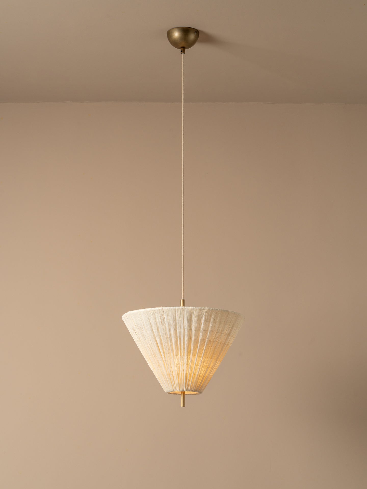 A modern pendant light illuminated with a pleated white conical shade and a brass ceiling mount, suspended against a minimalist beige wall and ceiling.