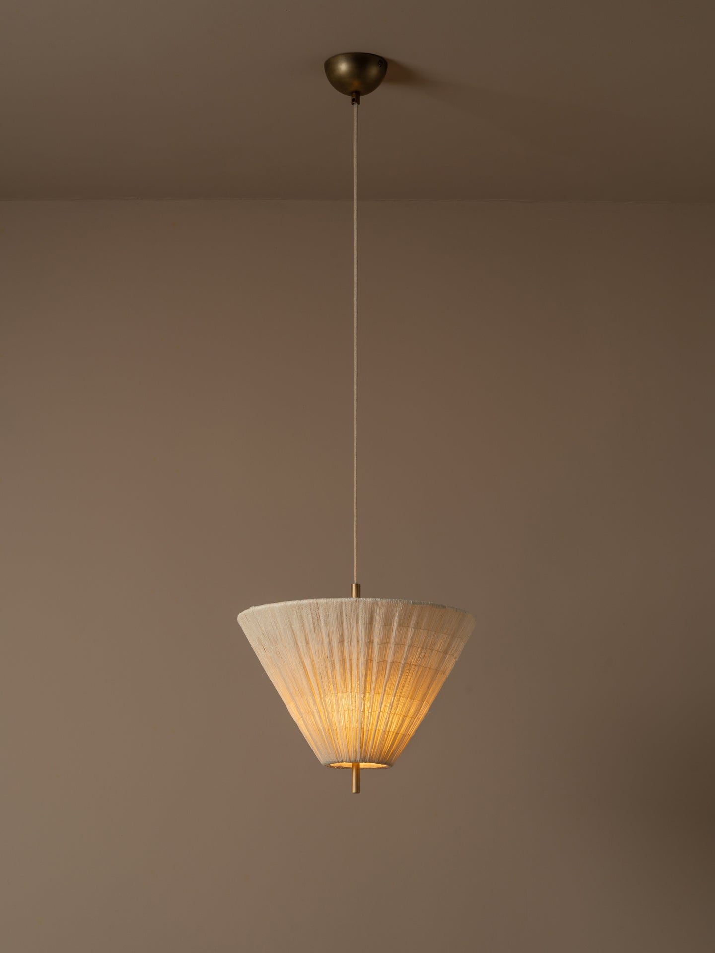 A modern pendant light illuminated with a pleated white conical shade and a brass ceiling mount, suspended against a minimalist beige wall and ceiling.