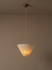 A modern pendant light illuminated with a pleated white conical shade and a brass ceiling mount, suspended against a minimalist beige wall and ceiling.