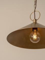 A close-up of a brass pendant light fixture with a wide, shallow conical shade and a glowing clear glass bulb, suspended against a plain beige background.