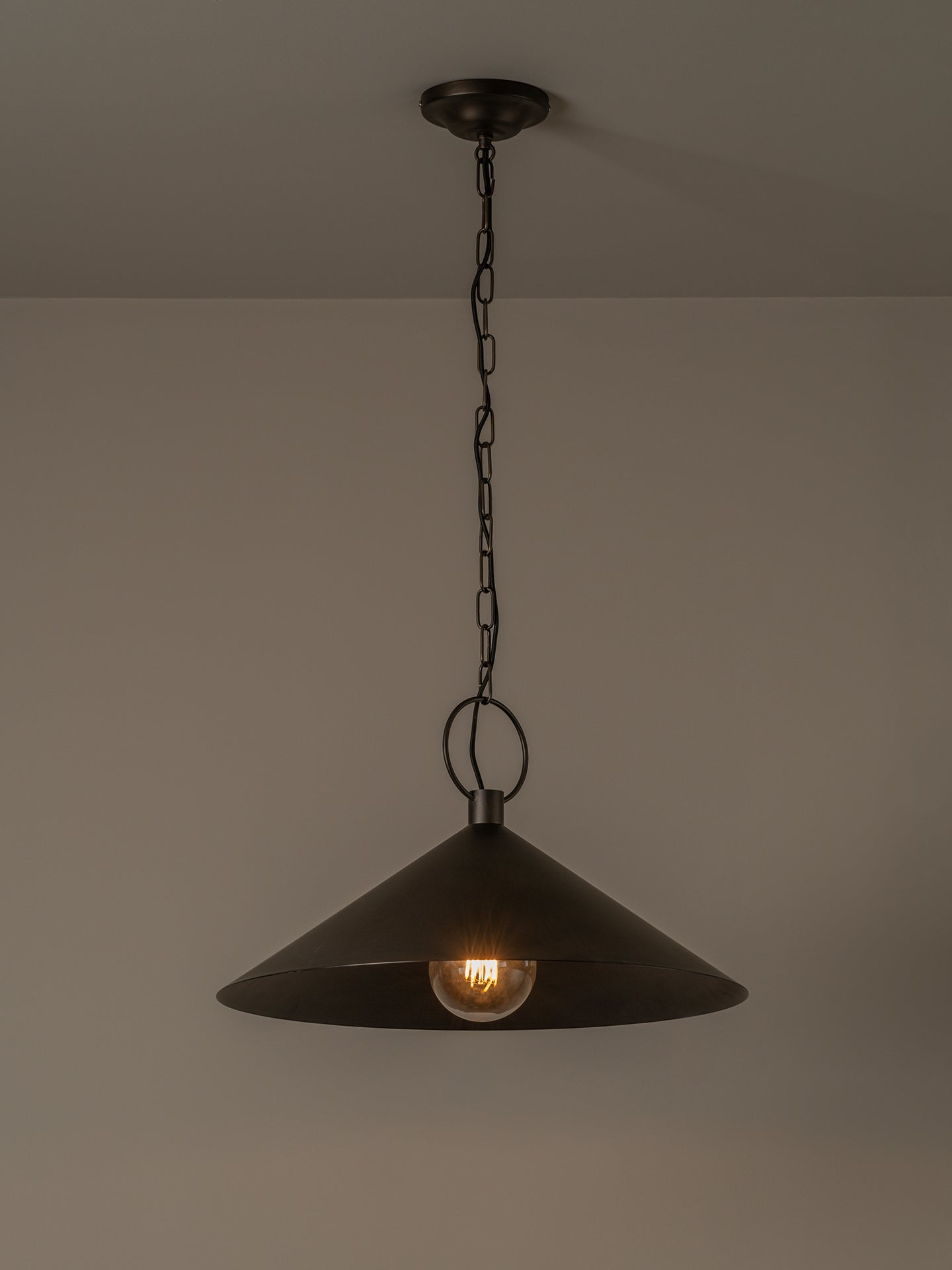 A dark metal pendant light with a wide conical shade hanging from a chain against a plain wall, with the light bulb turned on.