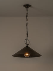 A dark metal pendant light with a wide conical shade hanging from a chain against a plain wall, with the light bulb turned on.