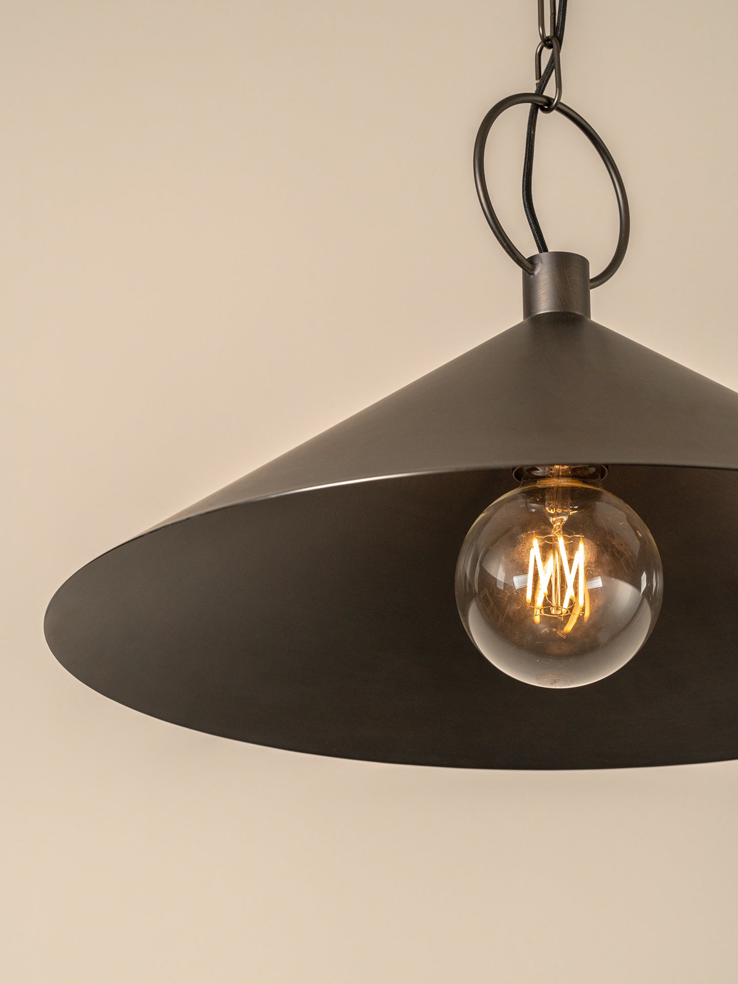A close-up of a dark metal pendant light with a wide conical shade and a visible glowing bulb, hanging against a plain beige wall.