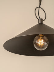 A close-up of a dark metal pendant light with a wide conical shade and a visible glowing bulb, hanging against a plain beige wall.