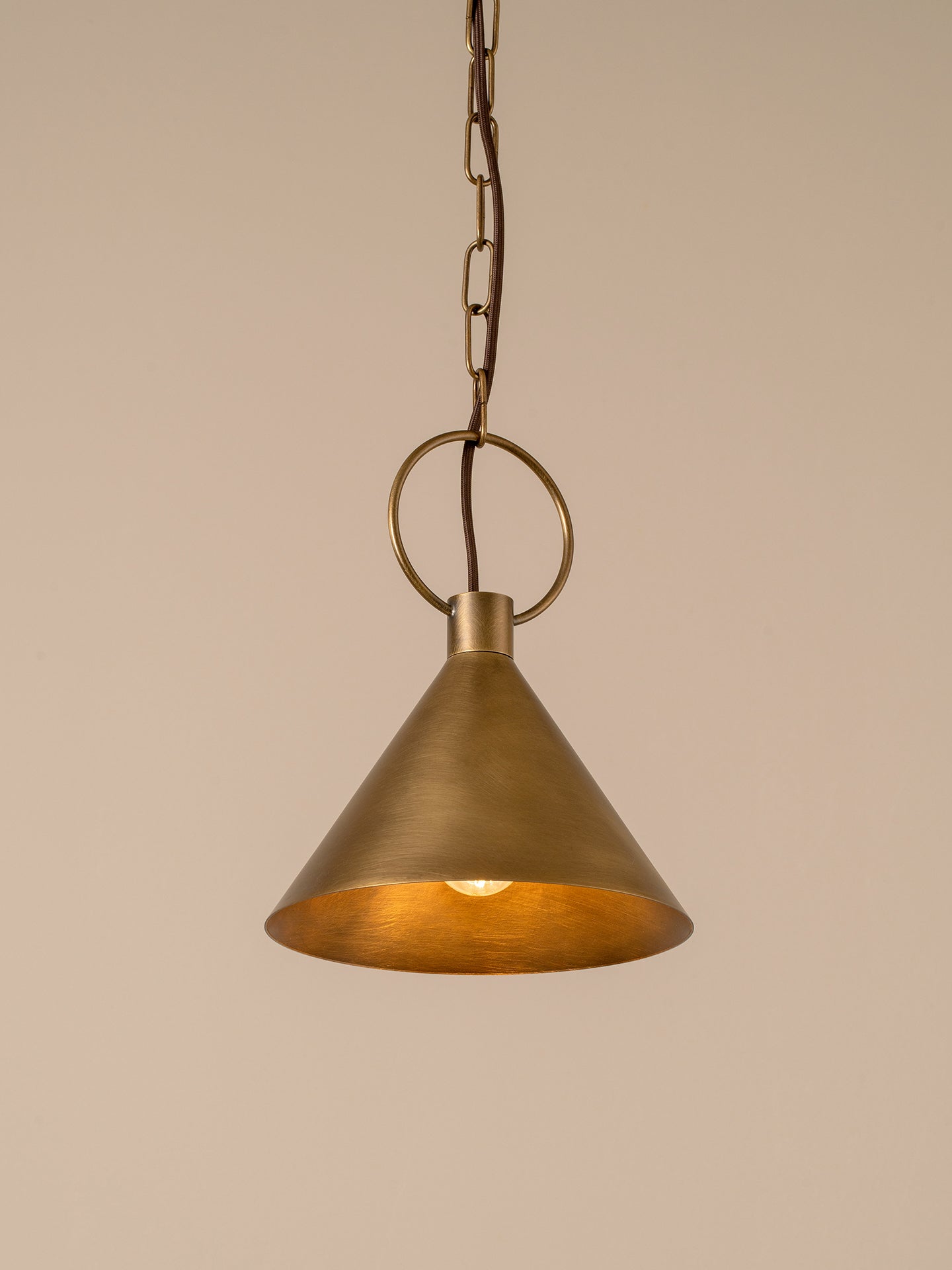 A single brass pendant light with a conical shade, hanging from a chain and ring against a solid beige background.