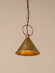 A single brass pendant light with a conical shade, hanging from a chain and ring against a solid beige background.