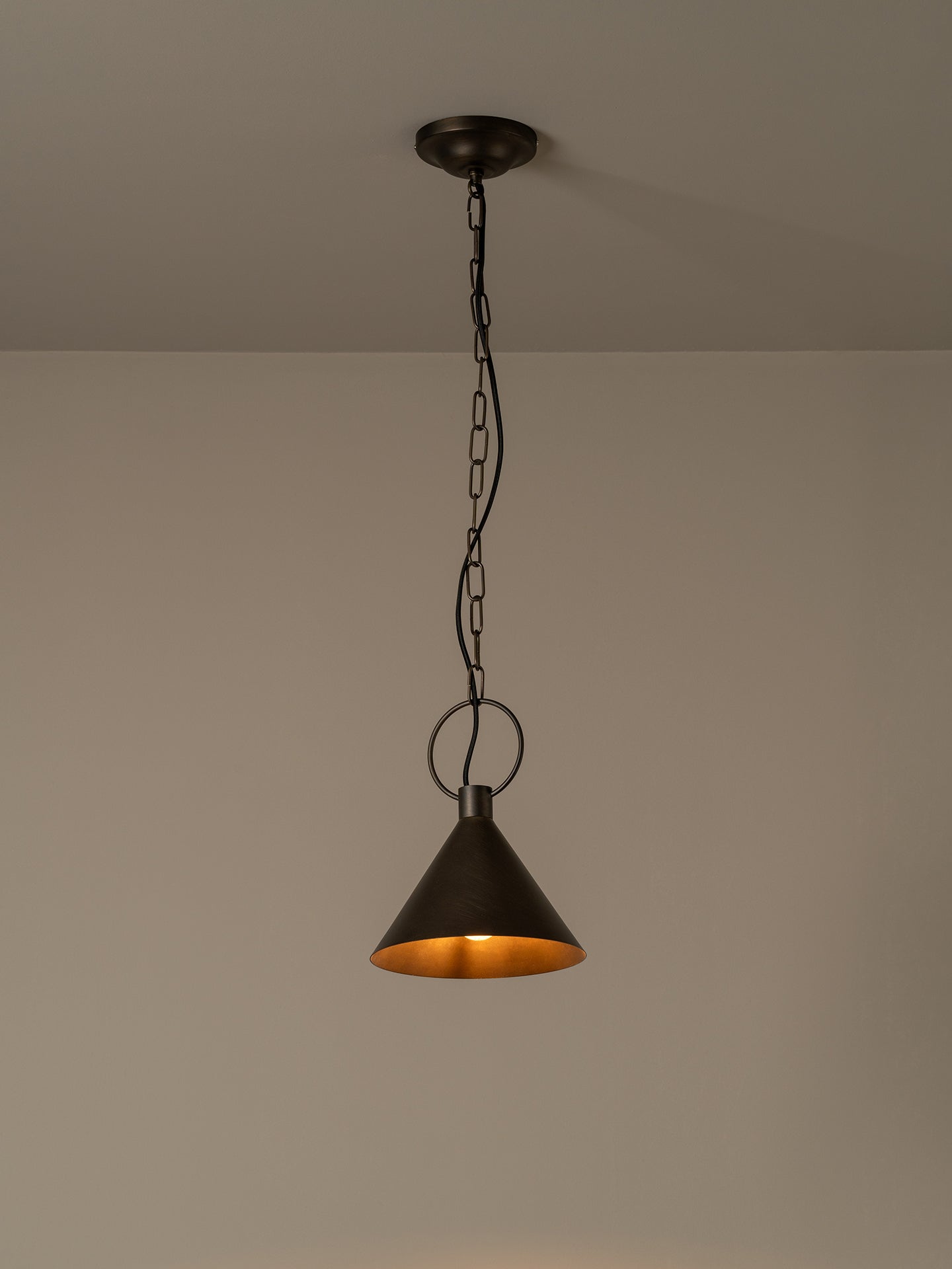 A dark metal pendant light with a conical shade and warm glowing interior, hanging from a chain against a plain beige wall and ceiling.