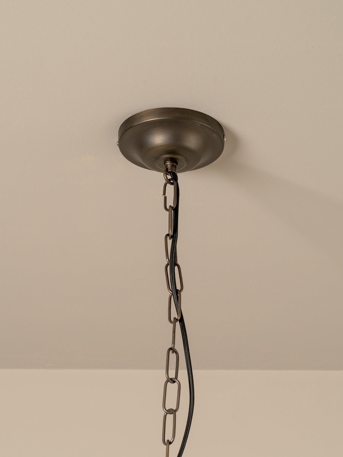 An industrial ceiling light fixture with a round, dark bronze ceiling canopy hanging from a heavy metal chain against a plain off-white wall.