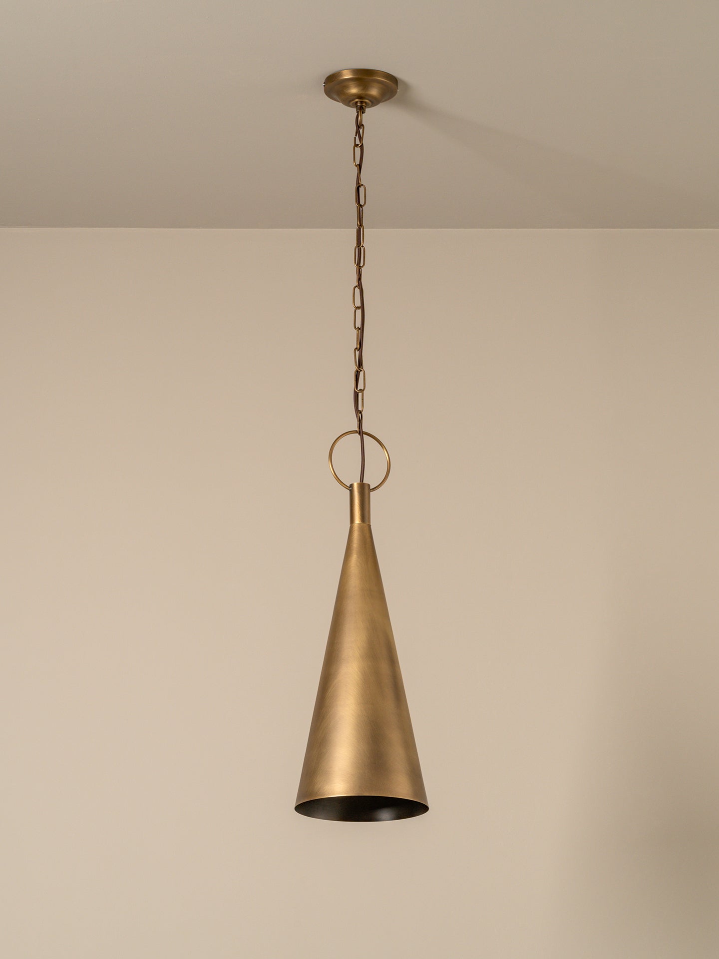 A brass cone pendant light hanging from a ceiling with a chain and ceiling mount, shown against a plain beige wall.