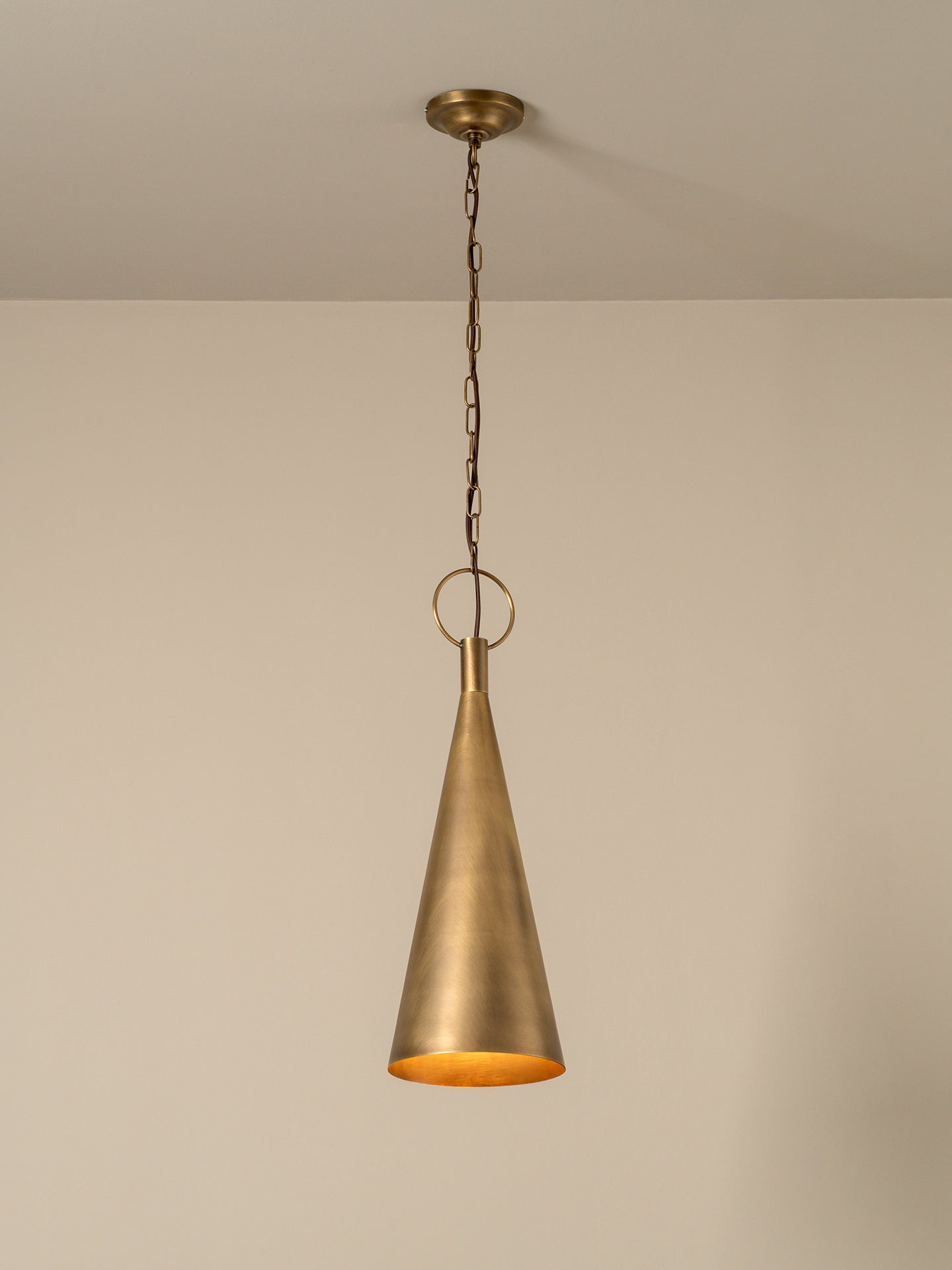A single brass pendant light with a conical shade hanging from a chain and ceiling canopy against a plain beige wall.
