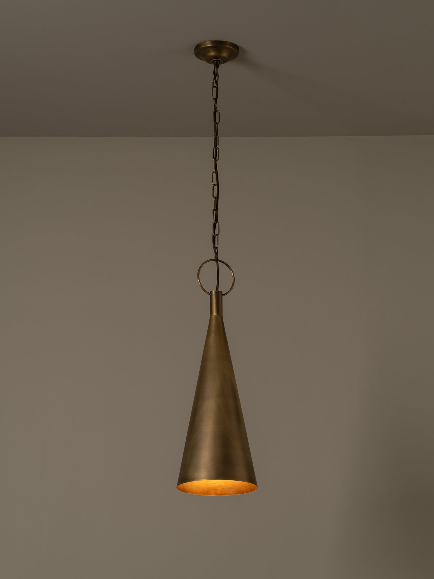 A single hanging pendant light with a tapered metal shade in a warm brass or bronze finish, suspended from a chain against a plain neutral wall.