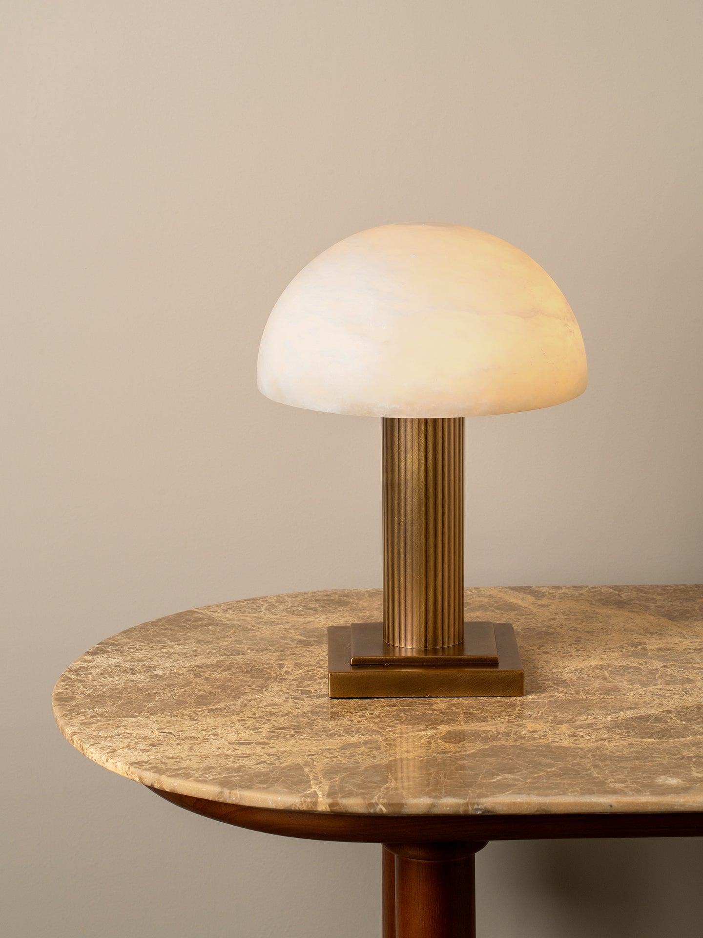 A mushroom-shaped table lamp with a glowing warm white dome shade and a fluted brass column base, displayed on a round marble-top table against a neutral beige wall.