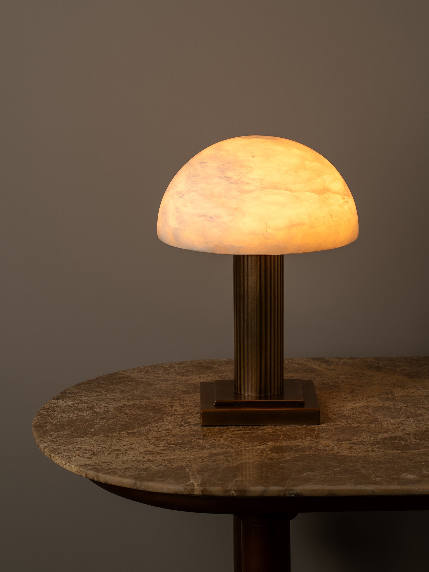 A mushroom-shaped table lamp with a glowing warm white marble-look dome shade and a ribbed metallic stem, displayed on a round stone-topped table against a dark gray background.