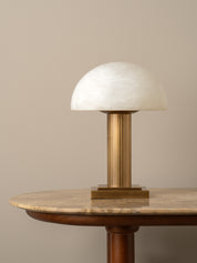 A modern table lamp with a white dome-shaped glass shade and a fluted brass column base, displayed on a round wooden table against a neutral beige background.