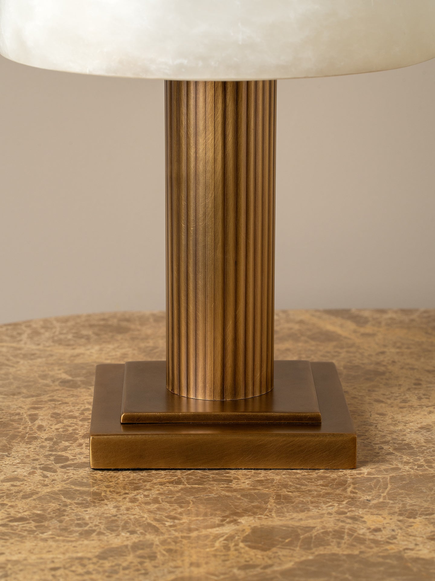 A close-up of a table lamp with a fluted metallic gold column base and a cream-colored oval shade, resting on a textured beige marble surface.