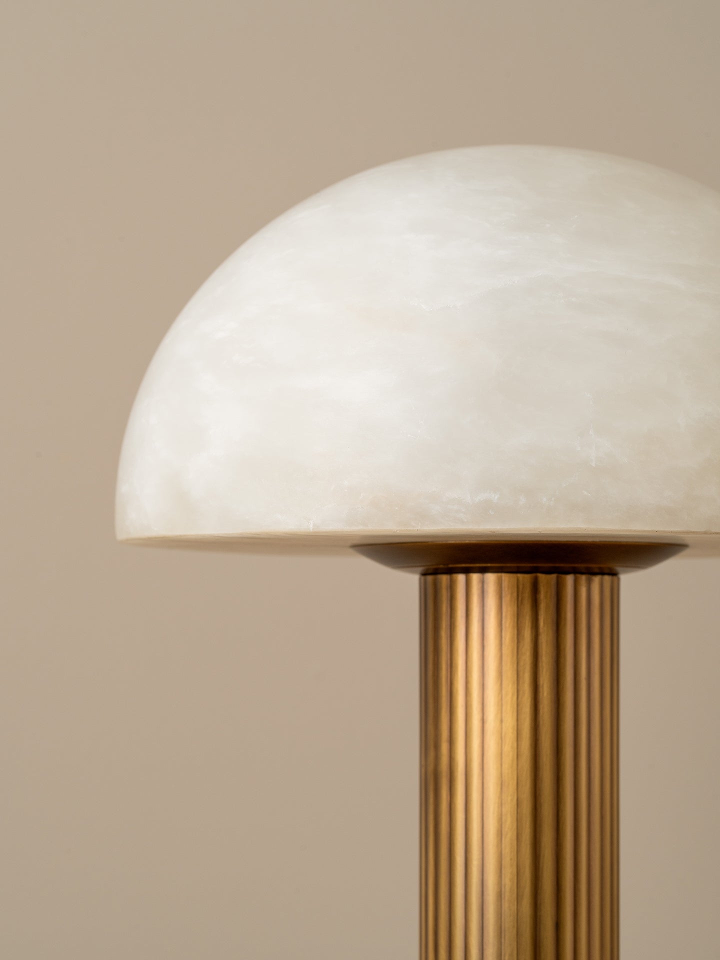 A close-up of a mushroom-shaped table lamp with a white marbled glass dome shade and a fluted brass column on a warm beige background.