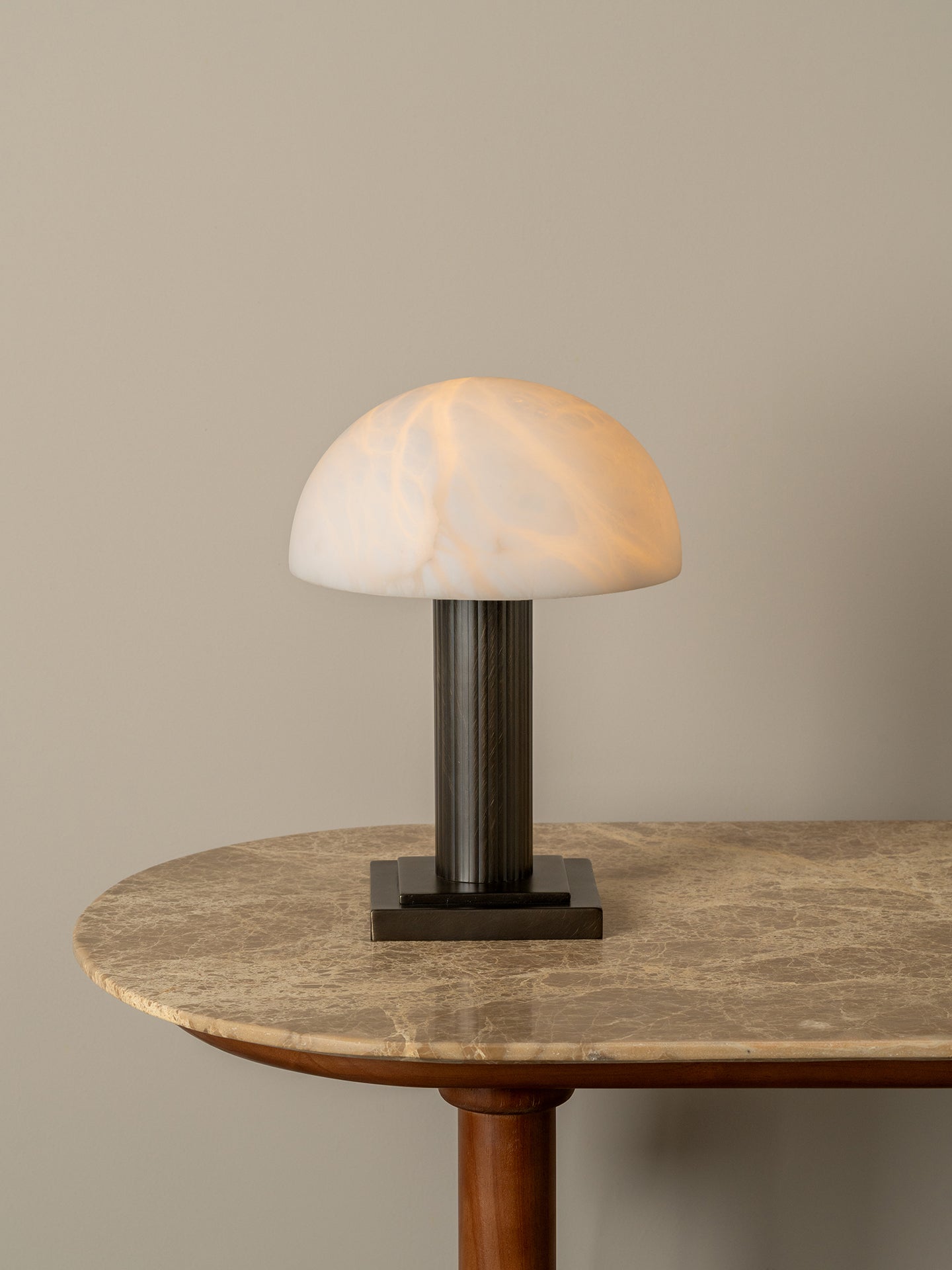 A mushroom-shaped table lamp with a glowing white marbled dome shade and a dark brushed metal stem, displayed on a round marble-topped table against a neutral beige wall.