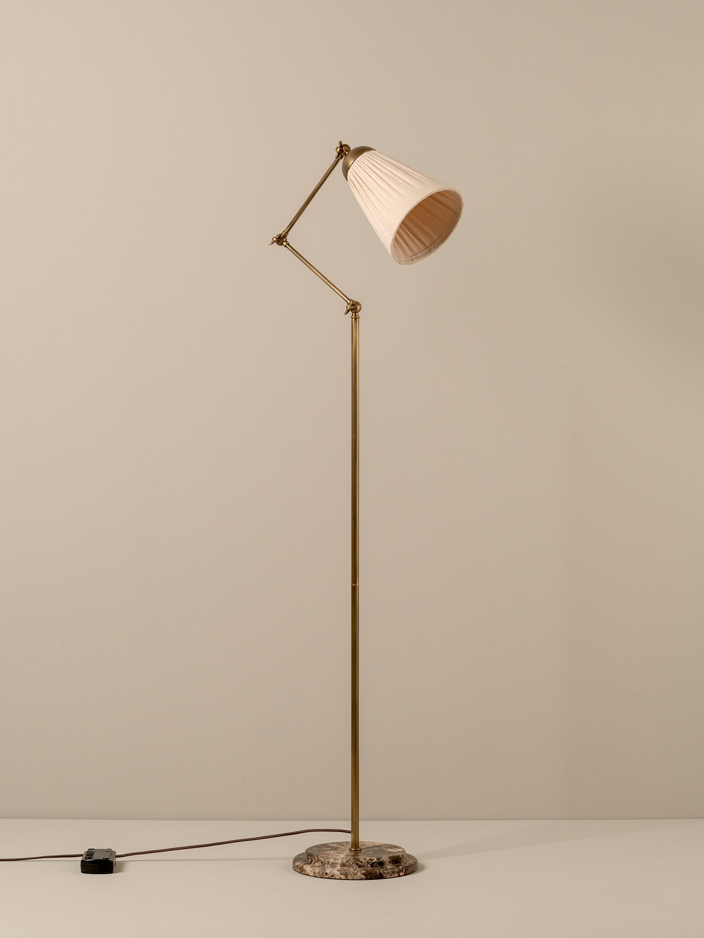 A mid-century modern floor lamp with a brass-colored metal frame, pleated off-white shade, and a round marble base, shown against a neutral beige background.