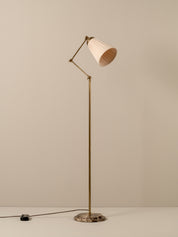 A mid-century modern floor lamp with a brass-colored metal frame, pleated off-white shade, and a round marble base, shown against a neutral beige background.