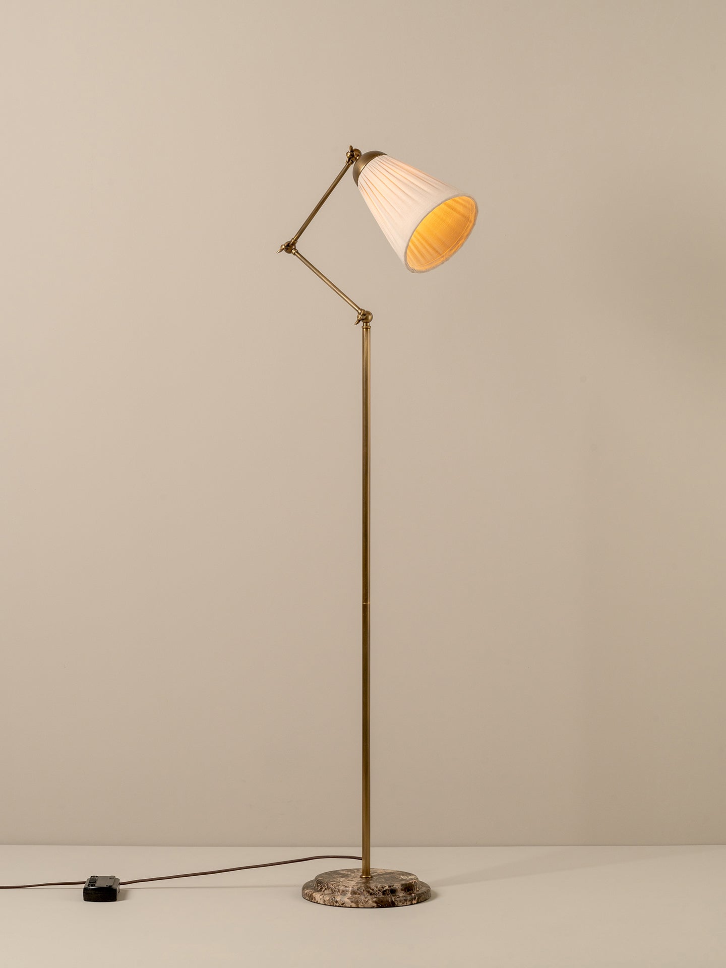A mid-century modern floor lamp with a brass-colored adjustable metal stand, a pleated white shade with a warm yellow-gold interior, and a round stone base, shown against a neutral beige background.